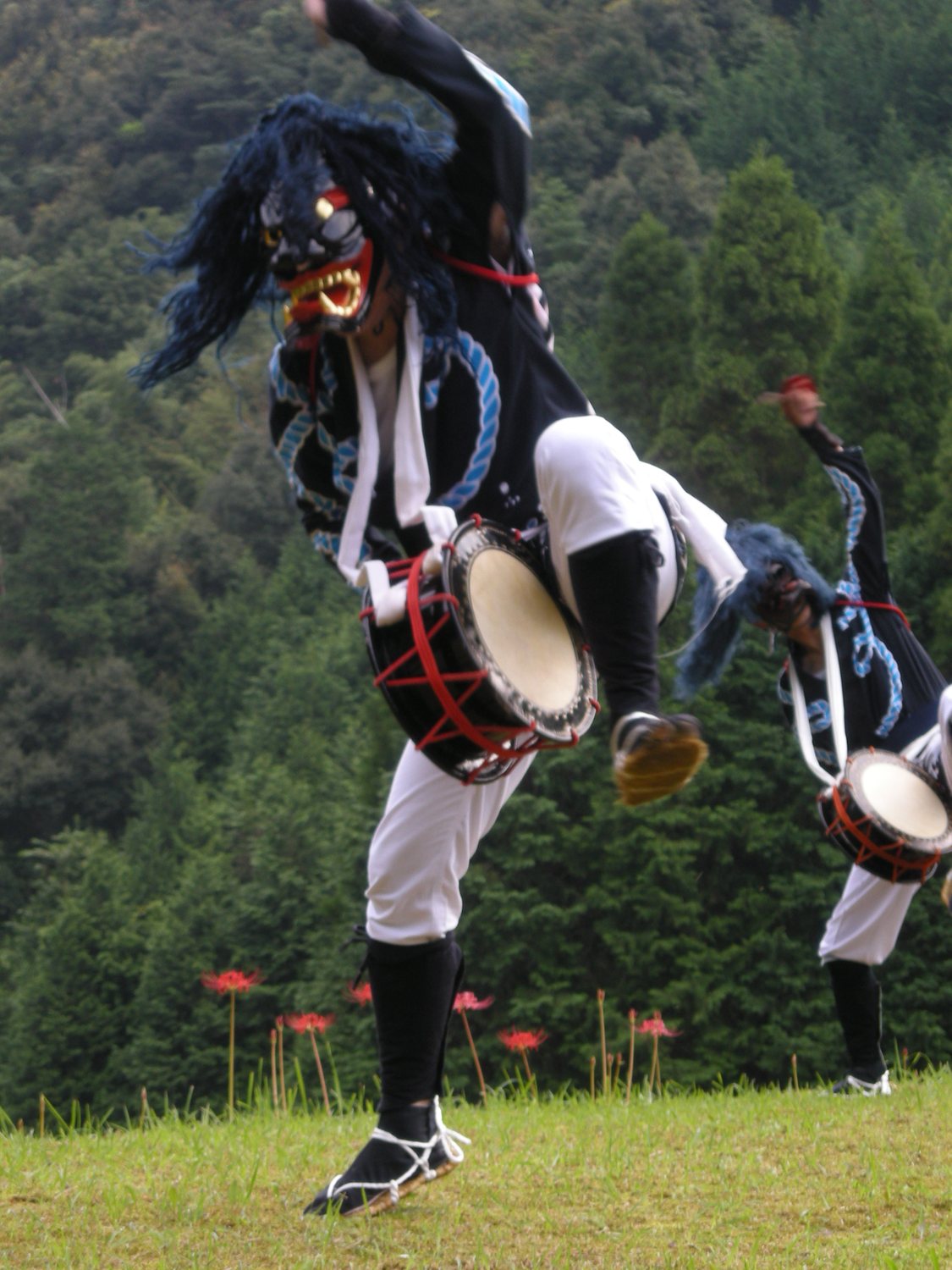 A men-buryu dancer mid-leap in a black lacquered oni mask and blue shaguma mane, waist drum swinging, red spider lilies in the grass behind