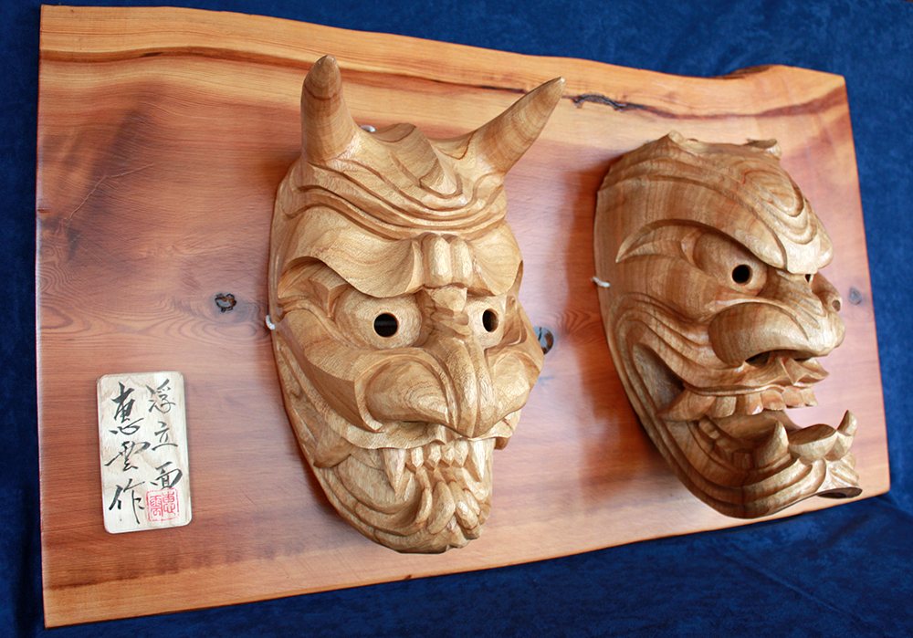 A pair of plain camphor furyu masks mounted on a live-edge cedar board with the carver's signed tag, on indigo cloth
