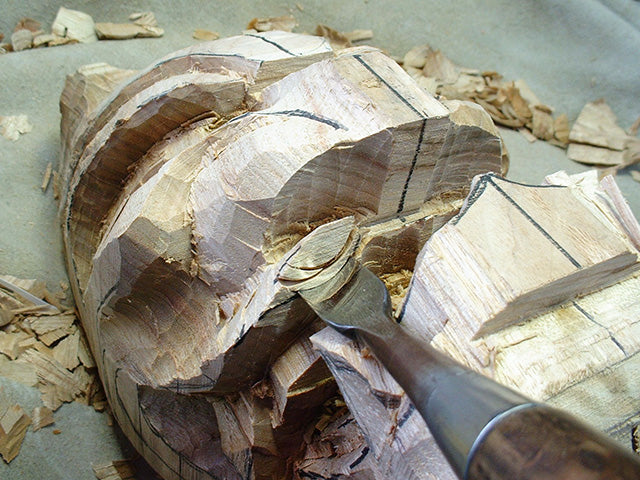 A chisel cutting the rough form of the oni face from the camphor block, shavings around it